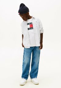 White and blue striped long-sleeve shirt over a white graphic t-shirt, loose blue jeans, and white sneakers, complemented by a navy beanie.