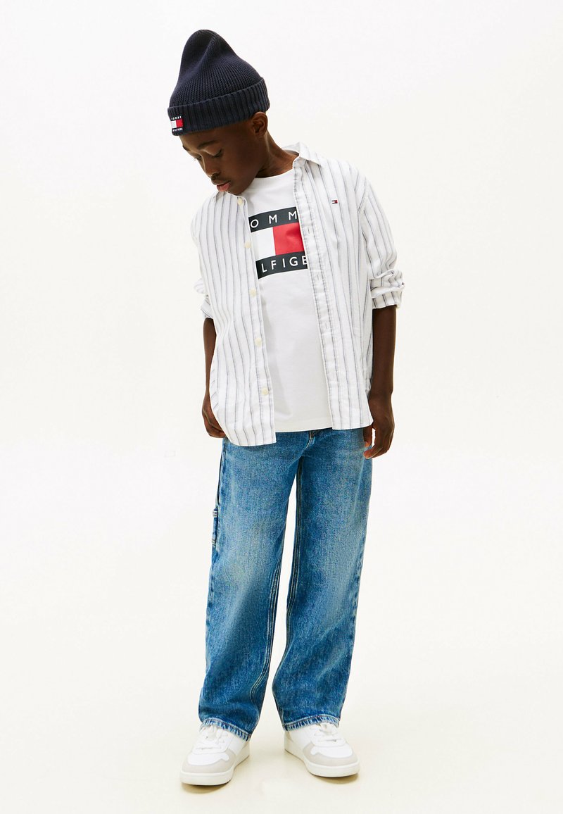 White and blue striped long-sleeve shirt over a white graphic t-shirt, loose blue jeans, and white sneakers, complemented by a navy beanie.