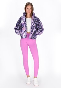 Kort jakke i sort, lilla og hvid marmor mønster, parret med pink ribbede leggings. Model iført hvide sko, smilende mod en ensfarvet baggrund.