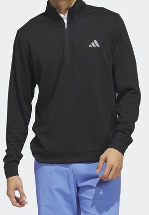 Mand iført sort quarter-zip pullover med Adidas-logo og lyseblå bukser, stående med afslappede arme mod en ensfarvet baggrund.