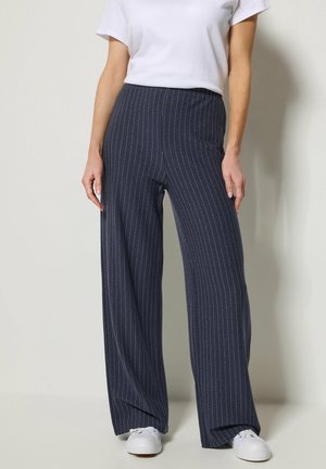 Frau trägt marineblaue, breitbeinige Nadelstreifenhose kombiniert mit weißen Turnschuhen und einem schlichten weißen Kurzarmshirt.