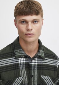 Cămașă din flanel în carouri verzi, cu dungi albe și negre, guler cu nasturi și buzunare pe piept. Țesătura pare groasă și texturată.