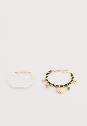 Deux bracelets sur surface blanche : un en perles blanches avec fermoir en or, un en chaîne d'or avec ruban vert et pendentifs en forme de cœur et de charme.
