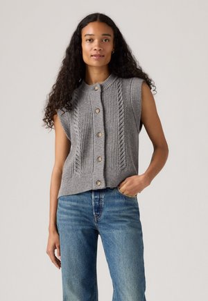 Gilet en tricot sans manches gris avec des motifs en relief, col rond et boutons en bois, associé à un jean taille haute bleu. Texture douce.