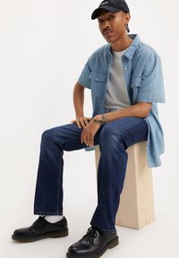 Camisa de mezclilla de manga corta de color azul claro sobre una camiseta blanca, jeans de color azul oscuro y zapatos negros, sentado sobre un bloque de madera con un fondo neutro.