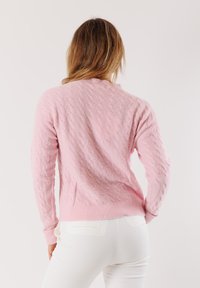 Persona con capelli castano chiaro che indossa un maglione rosa a trecce e pantaloni bianchi, di spalle contro uno sfondo chiaro e semplice.