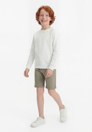 Ragazzo dai capelli rossi che sorride, camminando, indossando una maglietta bianca a maniche lunghe, pantaloncini verde oliva e scarpe da ginnastica bianche su uno sfondo bianco.