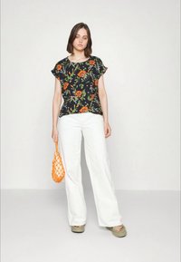 Top preto floral com design em laranja e verde, calças brancas de perna larga e uma bolsa de rede laranja texturizada. O modelo está de pé, descalço.