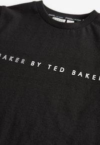 Črna bombažna majica s kratkimi rokavi, ki vsebuje belo besedilo "BAKER BY TED BAKER." Ima okroglo ovratnico in mehko, gladko teksturo.