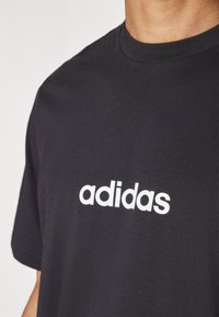 Juodas medvilnės marškinėliai trumpomis rankovėmis ir apvalia apykakle, su ryškiu baltu "adidas" logotipu ant krūtinės.