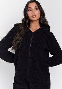 Mono de forro polar negro con cremallera frontal, capucha y tejido texturizado. Mangas largas con un ajuste relajado, adecuado para el uso casual.