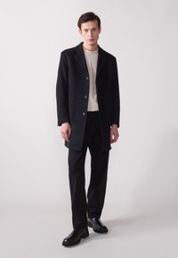 Abrigo de lana negro con cuello muesca, cinco botones y bolsillos laterales, usado sobre una camisa beige y pantalones negros, combinado con zapatos negros.