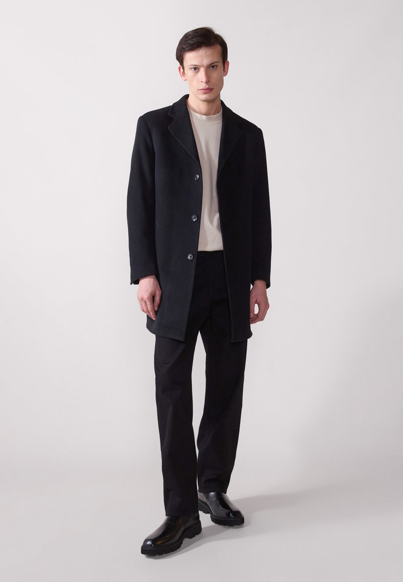 Abrigo de lana negro con cuello muesca, cinco botones y bolsillos laterales, usado sobre una camisa beige y pantalones negros, combinado con zapatos negros.