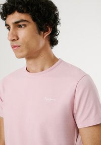 Tricou din bumbac de culoare roz deschis, cu guler rotund, mâneci scurte și logo-ul alb "Pepe Jeans" brodat pe piept. Textură netedă.