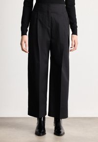 Άτομο που φοράει ψηλόμεσα, ευρύχωρα μαύρα παντελόνια σε ευρεία γραμμή, συνδυασμένα με μαύρα μποτάκια με μυτερή μύτη, στέκεται μπροστά σε απλό φόντο.