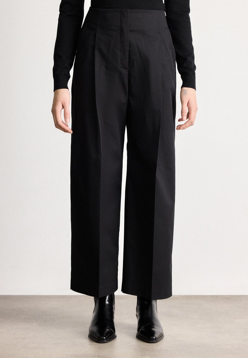 Άτομο που φοράει ψηλόμεσα, ευρύχωρα μαύρα παντελόνια σε ευρεία γραμμή, συνδυασμένα με μαύρα μποτάκια με μυτερή μύτη, στέκεται μπροστά σε απλό φόντο.