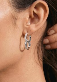 Pendientes de aro plateados con un diseño ondulado, que presentan pedrería clara a lo largo de un borde para añadir textura y detalle.