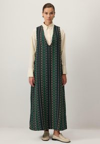 Rochie maxi neagră cu modele geometrice verzi și galbene, având un decolteu adânc în V și un croi lejer, purtată peste o bluză bej deschis.