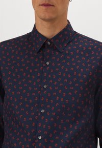 Man som bär en marinblå skjorta med knappar och röda abstrakta pilar mot en vit bakgrund. Han har också en silverfärgad hoopörhänge.