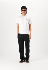 Biela bavlnená tričko s logom na ľavej hrudi, v kombinácii s čiernymi nohavicami vo voľnom strihu. Čierne tenisky a tmavé slnečné okuliare dopĺňajú celý outfit.