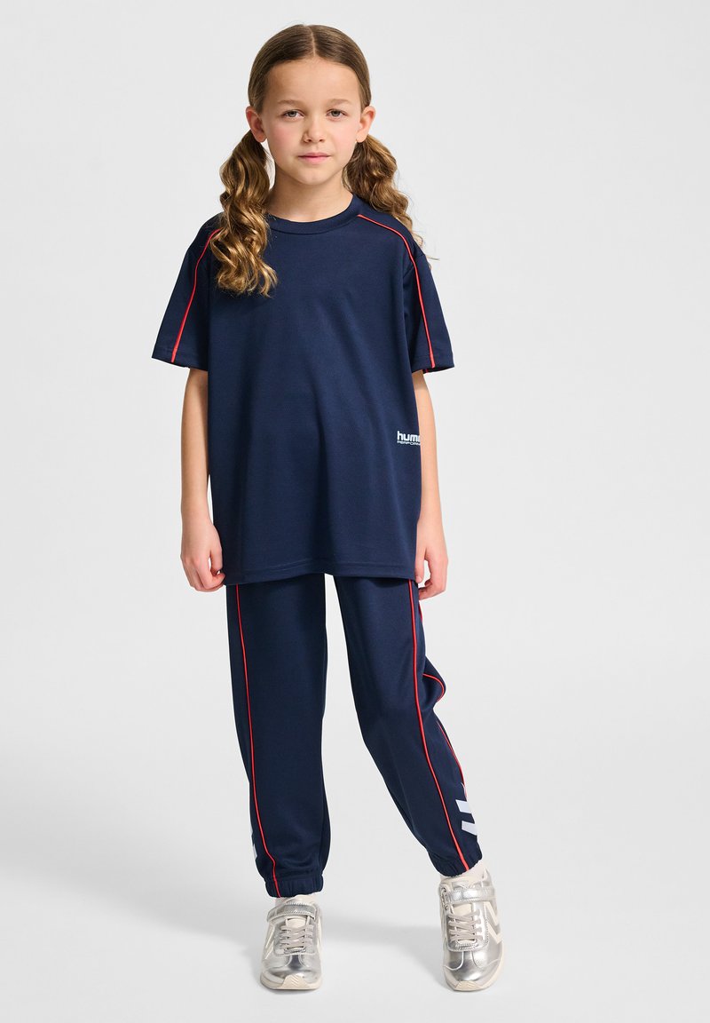 Marinblå träningsoveralls-set med röda detaljer; lös passform på t-shirten och byxorna med sidostrick. Texturerat material och logotypdetalj på tröjan. Silverfärgade sneakers.