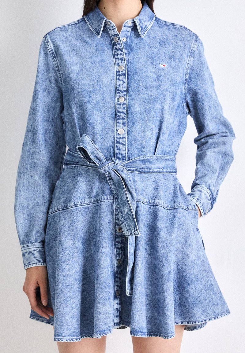 Robe en denim bleu clair avec des manches longues, un devant à boutons et une taille cintrée. Présente une jupe évasée et des détails de couture subtils.
