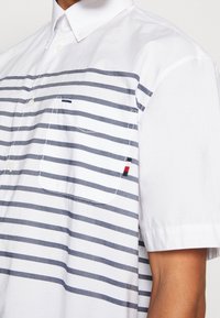 Vit kortärmad skjorta med marinblå horisontella ränder, knäppning med krage, vänster bröstficka och en liten röd, vit och blå detalj på fickan.