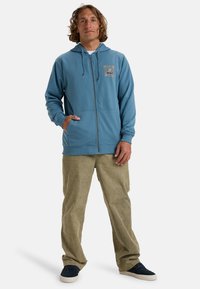 Light blue zip-up hoodie with drawstring hood and front pockets, worn over green corduroy pants. Features a small graphic on the chest.