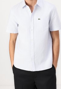 Lyhythihainen napillinen paita vaaleansinisissä ja valkoisissa raidoissa. Sisältää rintataskun ja vihreän Lacoste-logon. Puuvillakangas, rento istuvuus.