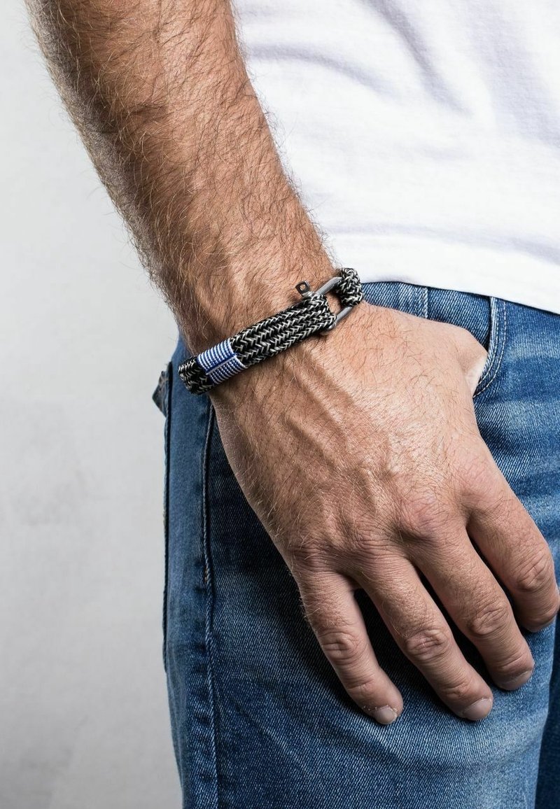 Main d'homme reposant sur la hanche, portant un jean bleu et une chemise blanche, avec un bracelet tissé noir et bleu au poignet.