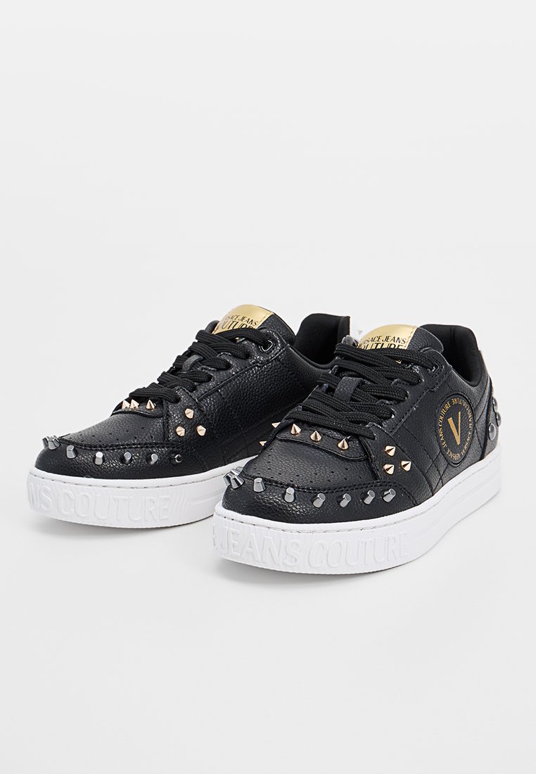 Zapatillas negras de piel con remaches metálicos, textura acolchada y suela de goma blanca. Cuenta con un parche de logo dorado en la lengüeta.