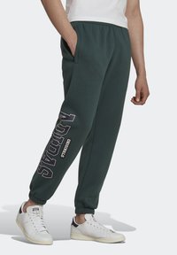 Zelené tepláky s elastickým pasem, bočními kapsami a nápisem "ADIDAS ORIGINALS" na levé noze. V kombinaci s bílými teniskami.