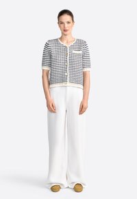 Veste courte texturée en noir et blanc à manches courtes avec des boutons dorés, associée à un pantalon blanc à jambes larges et des chaussures à enfiler marron.