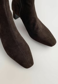 Donkere bruine suède enkellaarsjes met vierkante neuzen en zichtbare stiksels op een lichte achtergrond.