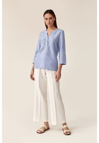 Top in lino azzurro chiaro con maniche a tre quarti e scollatura a V, con due bottoni sul davanti. Abbinato a pantaloni bianchi larghi e sandali beige.