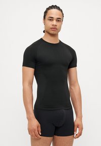 Schwarzes Sportshirt mit kurzen Ärmeln, enganliegendem Design, glatter Textur und dezentem Muster im Brustbereich, kombiniert mit schwarzen Shorts.