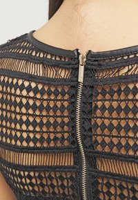 Prenda de encaje negro con un diseño geométrico que presenta recortes en forma de diamante. Un zipper dorado resalta la parte posterior, delineado con tela negra.