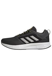 Zapatillas deportivas negras con una parte superior de malla texturizada, detalles plateados y cordones amarillos en contraste. Presentan una suela blanca acolchada.