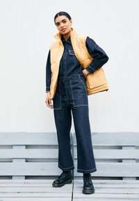 Chaleco acolchado amarillo sobre un mono de mezclilla oscura con piernas anchas. Botines negros con cordones. Fondo minimalista con palets de madera clara.