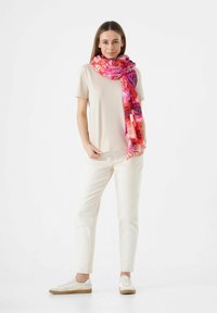 Foulard floral coloré avec des nuances de rose, rouge et violet drapé sur un haut beige. Le modèle porte un pantalon clair et des baskets blanches.