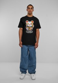 Mand med sort t-shirt med farverig kattegrafik og teksten "Velkommen God Formue," løse blå jeans og hvide sneakers står foran en grå baggrund.
