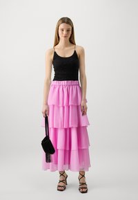 Svart spetstopp med justerbare stropper, parret med et lagdelt, ankel-lang rosa chiffon skjørt. Modellen holder en liten svart håndveske.