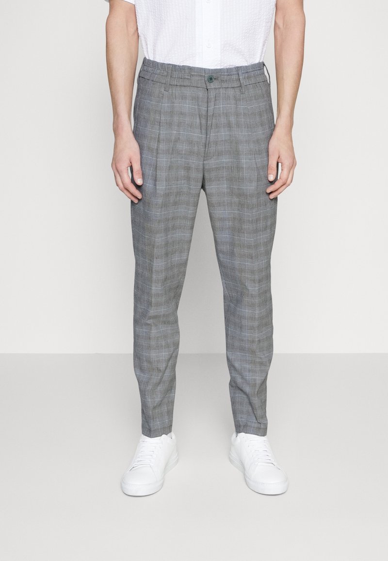 Homme portant un pantalon slim gris à carreaux avec des plis devant, des baskets blanches décontractées et une chemise blanche à manches courtes, debout devant un fond uni.