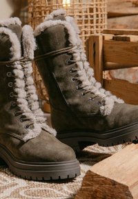 Zielono-oliwkowe zamszowe botki za kostkę z sztucznym futrem, posiadające metalowe oczka, sznurowaną konstrukcję oraz teksturowane gumowe podeszwy zapewniające dobrą przyczepność.