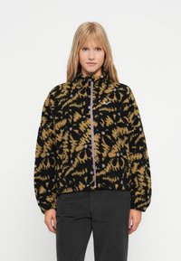 Mujer con una chaqueta de forro polar negra y mostaza con estampado floral, con cremallera frontal y cuello alto, combinada con pantalones negros frente a un fondo liso.