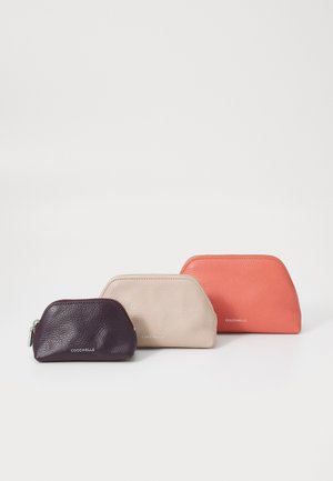 Three zippered pouches in dark purple, beige, and coral leather. They feature rounded tops and embossed branding on the front.