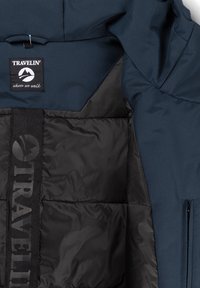 Marineblauwe gewatteerde jas met zwart doorgestikte voering, label "Travelin' where we walk" en zwarte gebrandmerkte binnenbanddetails.