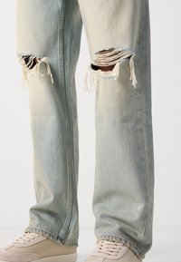 Vaqueros desgastados de color azul claro con cortes rasgados en las rodillas, ajuste tapered y una textura deslavada, complementados con zapatillas beige.