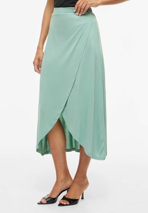 Frau trägt einen hellgrünen, Wickel-Midirock, kombiniert mit schwarzen, offenen Sandalen mit Absatz vor einem weißen Hintergrund.