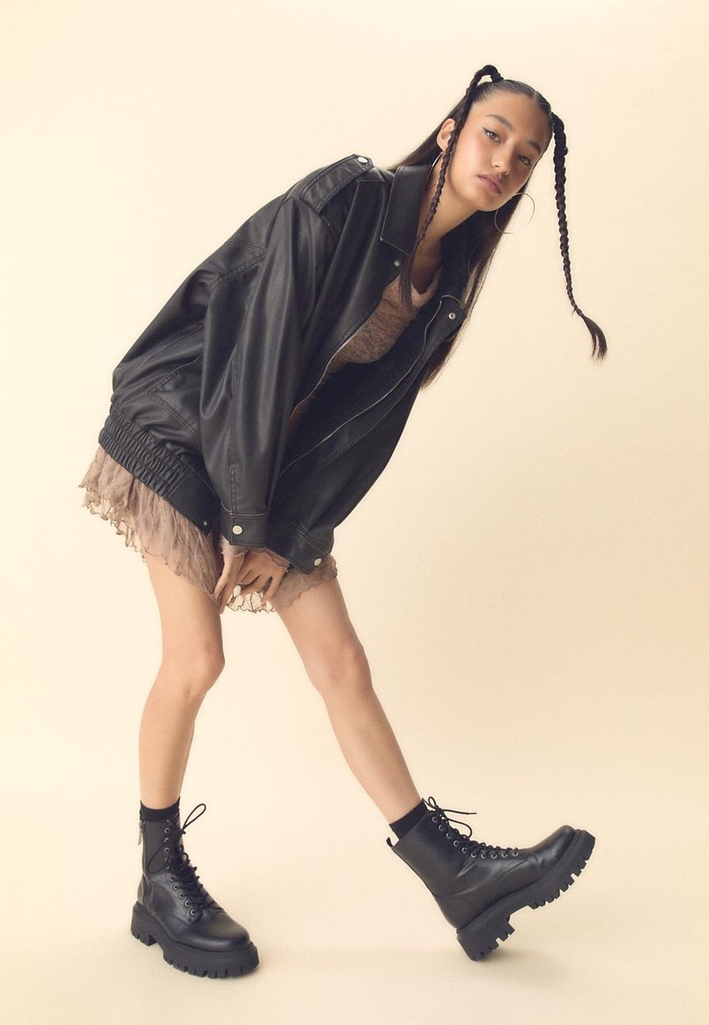Giacca oversized nera in pelle con vita elasticizzata, indossata sopra un vestito beige trasparente con orlo a volant. La modella indossa stivali neri chunky con lacci.